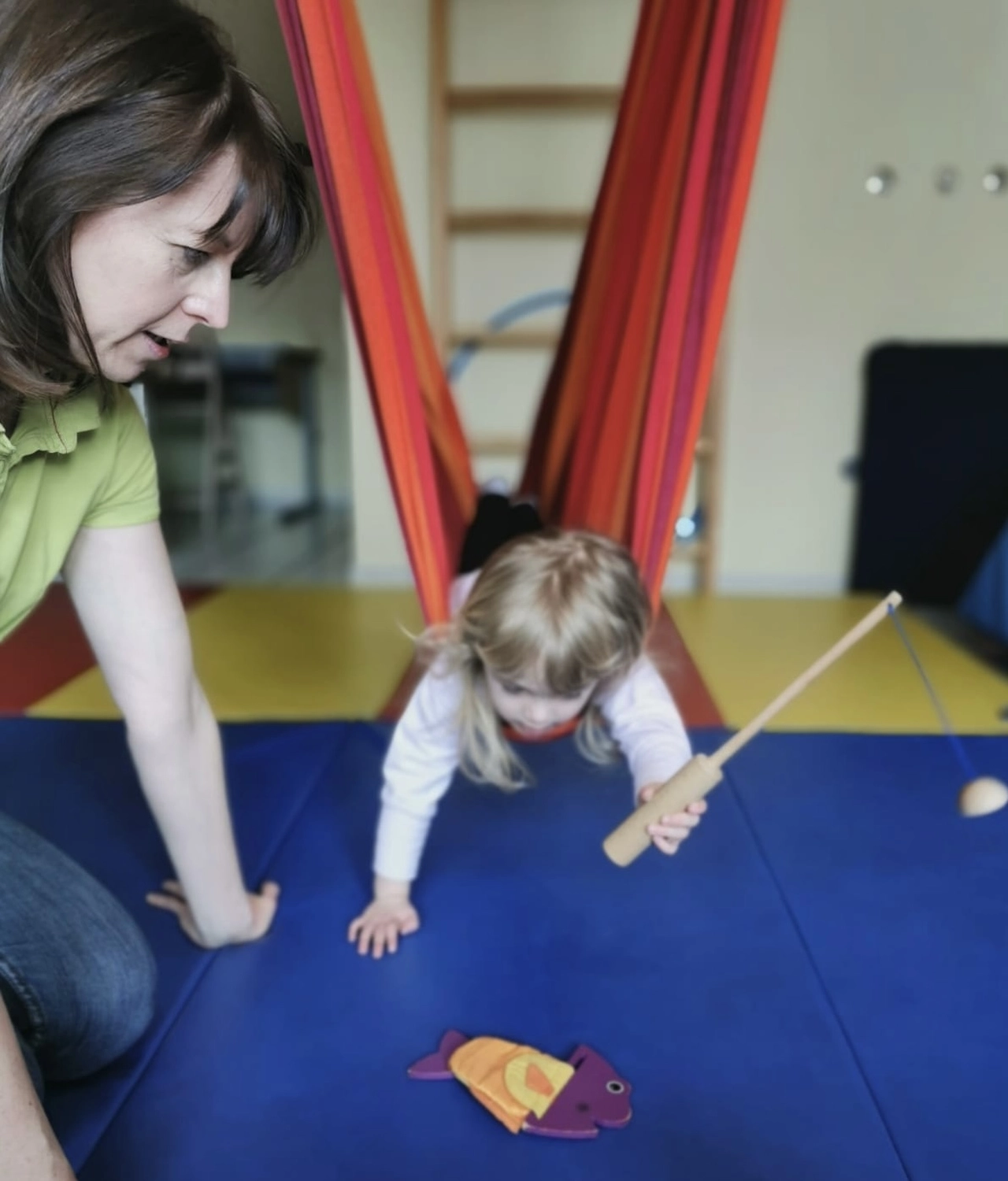 Praxis für Ergotherapie - medizinisch-therapeutische Behandlung - Eine Frau übt mit einem kleinen Kind, das in einer Schaukel hängt, das Fangen eines Spielzeugs.